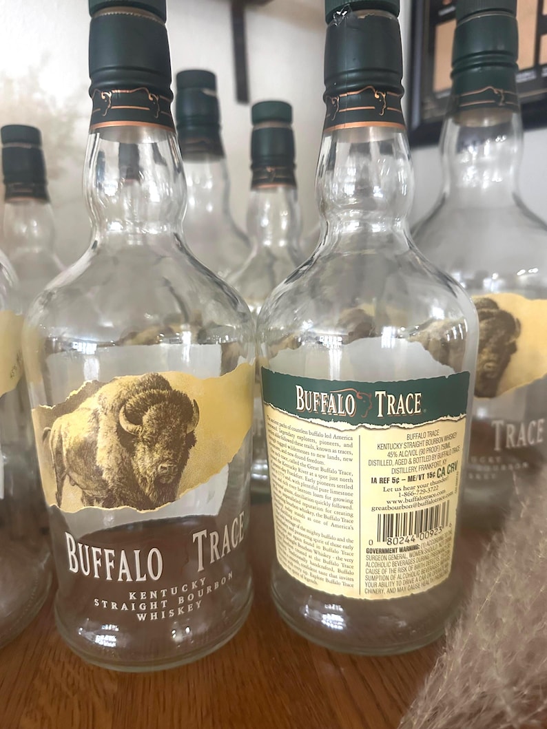 May include: Several empty Buffalo Trace Kentucky Straight Bourbon Whiskey bottles. The clear glass bottles have green and gold labels with the brand name and a bison illustration. The bottles are arranged on a wooden surface.
