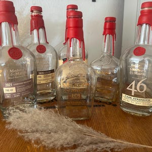Pode incluir: V&aacute;rias garrafas de whisky Maker's Mark de vidro transparente com selos de cera vermelha. Algumas garrafas t&ecirc;m r&oacute;tulos indicando edi&ccedil;&otilde;es limitadas e teor alco&oacute;lico. As garrafas est&atilde;o dispostas sobre uma superf&iacute;cie de madeira.