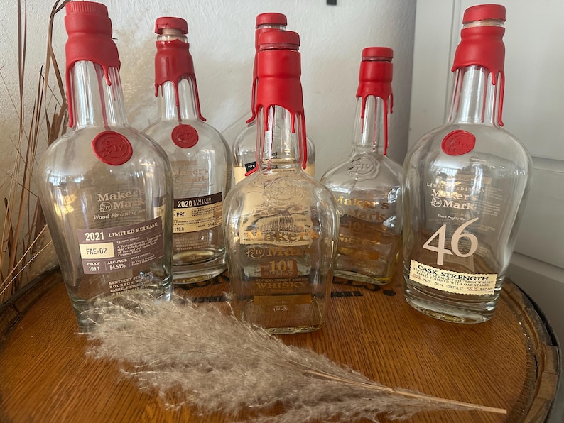Pode incluir: V&aacute;rias garrafas de whisky Maker's Mark de vidro transparente com selos de cera vermelha. Algumas garrafas t&ecirc;m r&oacute;tulos indicando edi&ccedil;&otilde;es limitadas e teor alco&oacute;lico. As garrafas est&atilde;o dispostas sobre uma superf&iacute;cie de madeira.