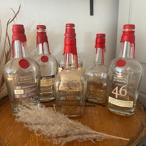 Pode incluir: Uma cole&ccedil;&atilde;o de garrafas de u&iacute;sque Maker's Mark com selos de cera vermelha. As garrafas, com r&oacute;tulos como "2021 Limited Release" e "46 Cask Strength", est&atilde;o dispostas em um barril de madeira. Uma pena decorativa adiciona um toque r&uacute;stico.