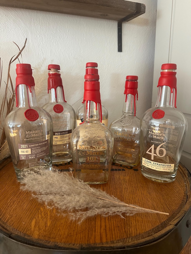 Pode incluir: Uma cole&ccedil;&atilde;o de garrafas de u&iacute;sque Maker's Mark com selos de cera vermelha. As garrafas, com r&oacute;tulos como "2021 Limited Release" e "46 Cask Strength", est&atilde;o dispostas em um barril de madeira. Uma pena decorativa adiciona um toque r&uacute;stico.