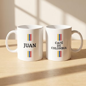 May include: Two white ceramic coffee mugs. One mug has the name "JUAN" printed in black with a vertical stripe of yellow, blue, and red. The other mug says "CAFÉ DE COLOMBIA" in black with a similar stripe.