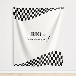 May include: White wall hanging with black and white checkered flag design at the top and bottom. The text "RIO IS... Formula 1" is printed in black script in the center of the wall hanging.