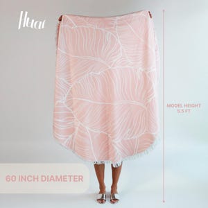 May include: A pink round beach towel with a white leaf pattern and fringe. The towel is 60 inches in diameter. A model is holding the towel in front of her, and the model is 5.5 feet tall.