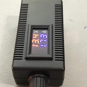 May include: A black electronic device with a digital display showing voltage and amperage readings. The display has red and blue illuminated numbers. A knob is located at the bottom of the device.