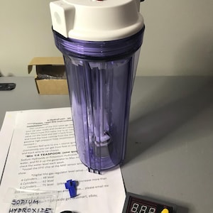 May include: A clear, cylindrical water filter housing with a white top and purple tinted body. The top has two metal fittings. A digital display, a bag of Sodium Hydroxide, and instructions are also present.
