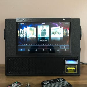 May include: A black digital jukebox with a touchscreen display showing album covers, a speaker grille, and a bill acceptor slot labeled INSERT. Included are a small remote control, a set of keys, and a compact wireless keyboard with a trackpad.