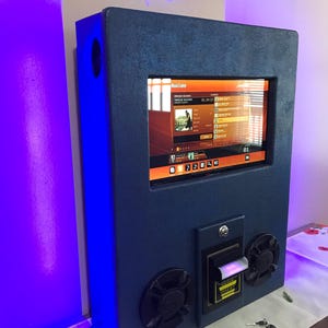 May include: A blue karaoke machine with a touch screen display showing the song "Everybody Dance" by the artist "Everybody Dance". The machine has a slot for inserting money and two speakers on the front.