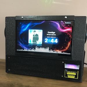 Puede incluir: Una gramola digital negra con una pantalla rectangular que muestra la canción Contigo de Enrique Iglesias, un temporizador de 2:44 y un gráfico de ondas sonoras. La unidad cuenta con una rejilla de altavoz frontal y un aceptador de billetes.