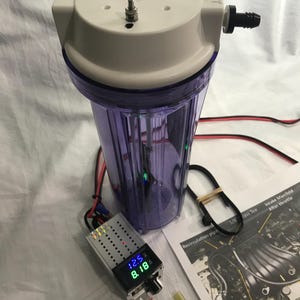 Könnte beinhalten: Ein transparentes, violettes, zylindrisches Wasserfiltergehäuse mit weißer Kappe, zwei Metallbolzen und einem schwarzen Schlauchanschluss. Ein digitales Anzeigemodul zeigt 12.5 und 8.18 in Blau und Grün, dazu ein Beutel mit SODIUM HYDROXIDE.