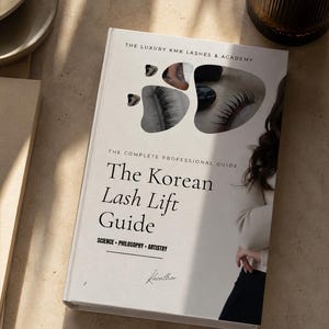 May include: A white hardcover book titled The Korean Lash Lift Guide, The Complete Professional Guide by The Luxury KMK Lashes & Academy. The cover features black text, the subtitle Science - Philosophy - Artistry, and abstract cutouts showing eyelashes.