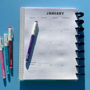 May include: A planner with a January calendar page, featuring a black spiral binding and a clear cover. Four colored pens, red, blue, and purple, are next to the planner. The background is a solid light blue.