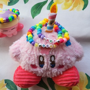 Op de afbeelding: Een roze pluche speelgoed met een regenboog kralenketting met de tekst "PARTY POOB". Een klein roze beeldje met een paddenstoelhoed en een blauwe knoop met een varenontwerp zijn ook aanwezig. Er staat een kaars op het pluche speelgoed.