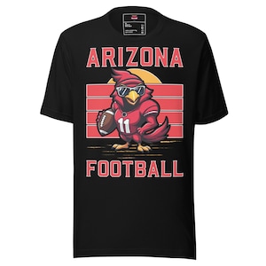 Op de afbeelding: Zwart t-shirt met een cartoon kardinaal met een zonnebril en een voetbaluniform, met een voetbal. Het shirt heeft de woorden "ARIZONA FOOTBALL" in rode letters. De kardinaal heeft nummer 11 op zijn uniform.