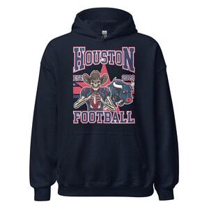 Op de afbeelding: Marineblauwe hoodie met een afbeelding van een skelet met een cowboyhoed en een voetbal. Het ontwerp bevat de tekst "Houston Football" en een illustratie van een stier. De hoodie is een pullover-model.