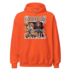 Puede incluir: Sudadera con capucha naranja con un gráfico que presenta un tigre, un jugador de fútbol esqueleto y las palabras "Cincinnati Football" en blanco y naranja. La sudadera tiene mangas largas y una capucha.