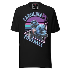 May include: Black t-shirt featuring a stylized illustration of a panther holding a football. The design includes the words "Carolina Football" in a retro font, with a purple and blue sunset background. A football player is also in the design.