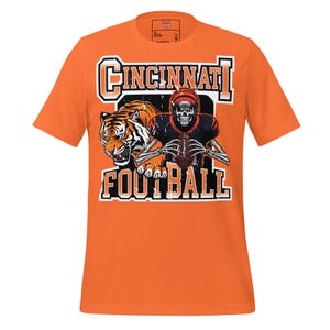 Puede incluir: Camiseta naranja con un gráfico de un tigre y un jugador de fútbol esqueleto. El texto "Cincinnati Football" se muestra encima del gráfico en una fuente en negrita y con contorno. La camiseta tiene mangas cortas y cuello redondo.