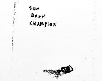 May include: A white rectangular print featuring the black hand-drawn text STAY DOWN CHAMPION in the upper centre. Below the text, a small, textured black graphic resembling a fallen object sits on the otherwise blank white surface.
