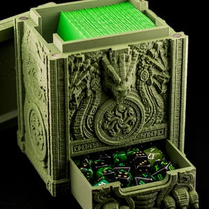 May include: A sage green Hydra Temple Commander deck box with a textured, sculpted exterior featuring a multi-headed serpent design. The box includes a magnetic lid, a pull-out drawer for dice, and an open top compartment holding green cards.