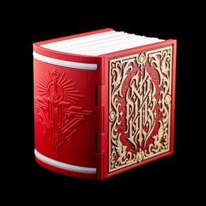 May include: A decorative red book with ornate gold filigree on the cover. The spine and front cover display detailed geometric designs. The pages are white, and the edges have white trim. The book is set against a black background.