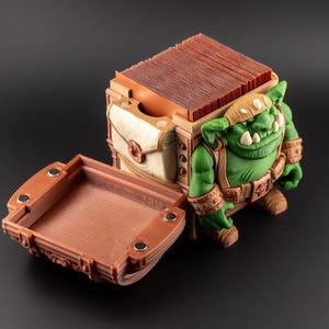 May include: A detailed, open treasure chest with a green goblin figure. The chest is brown with a white and tan pouch. The goblin has a green complexion, a brown headband, and wears a brown belt and wristbands. The chest's tray is open.