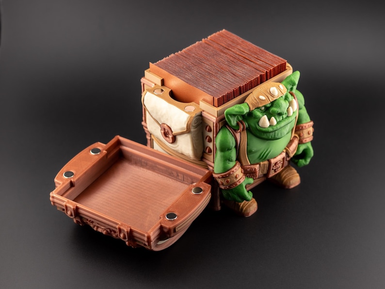 May include: A detailed, open treasure chest with a green goblin figure. The chest is brown with a white and tan pouch. The goblin has a green complexion, a brown headband, and wears a brown belt and wristbands. The chest's tray is open.