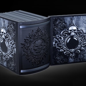 May include: A black book with a skull and ornate design on the cover. The book is open and the pages are visible. The book is made of a hard material and has a textured surface.