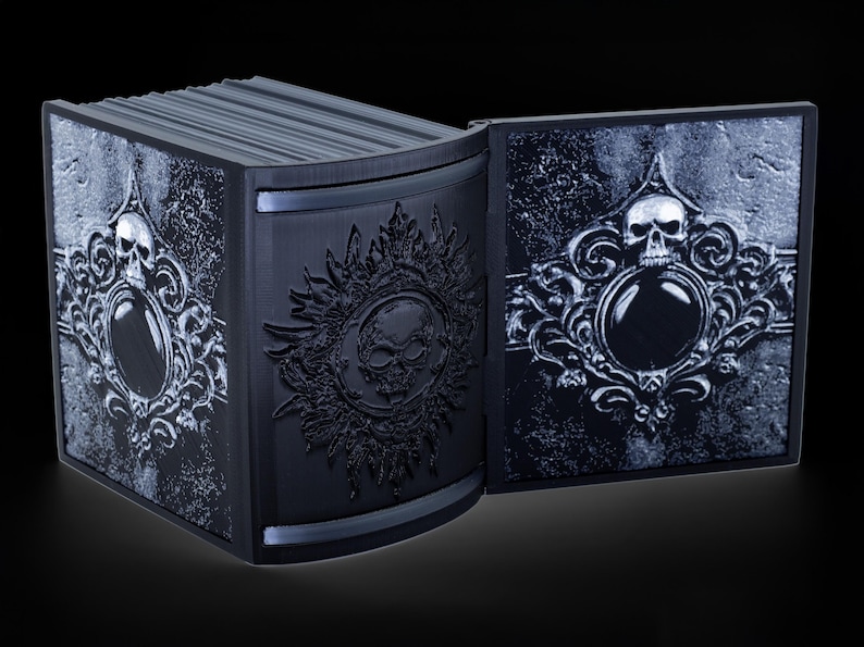May include: A black book with a skull and ornate design on the cover. The book is open and the pages are visible. The book is made of a hard material and has a textured surface.