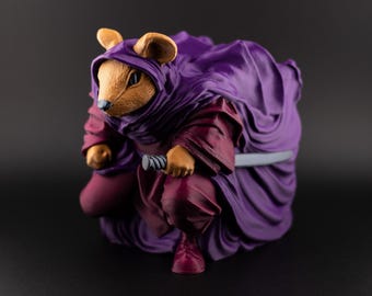 May include: A figurine of a mouse dressed in a purple hooded cloak and burgundy clothing, holding a black object. The mouse is in a crouching position, with its ears and face visible. The background is black.