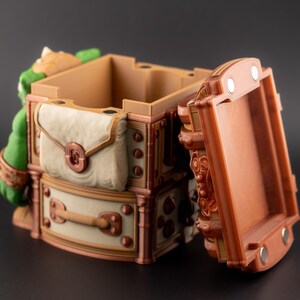 May include: A detailed, open treasure chest with a hinged lid. The chest is primarily brown with cream and copper accents, featuring a satchel and decorative hardware. A green figure is visible on the side.