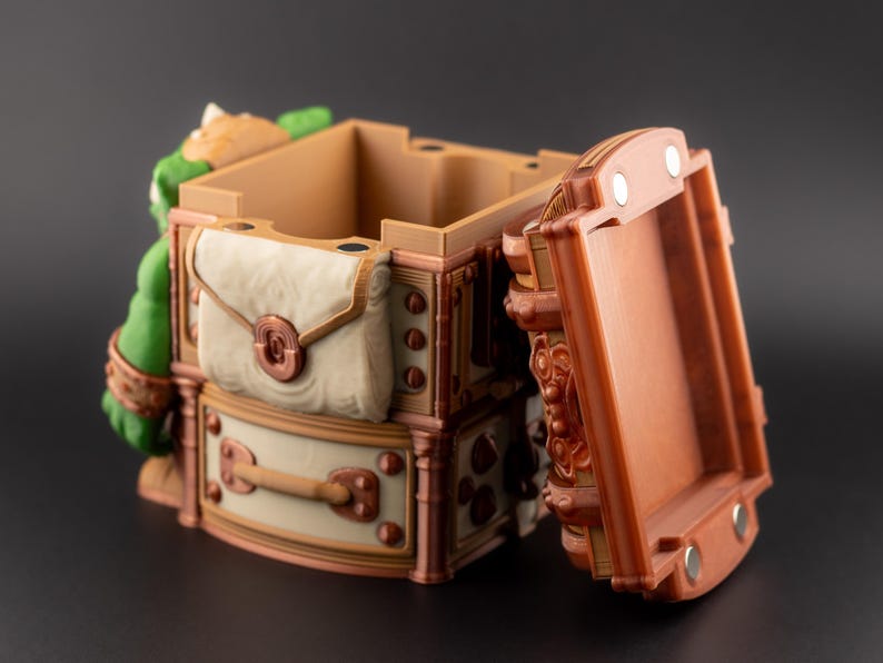 May include: A detailed, open treasure chest with a hinged lid. The chest is primarily brown with cream and copper accents, featuring a satchel and decorative hardware. A green figure is visible on the side.