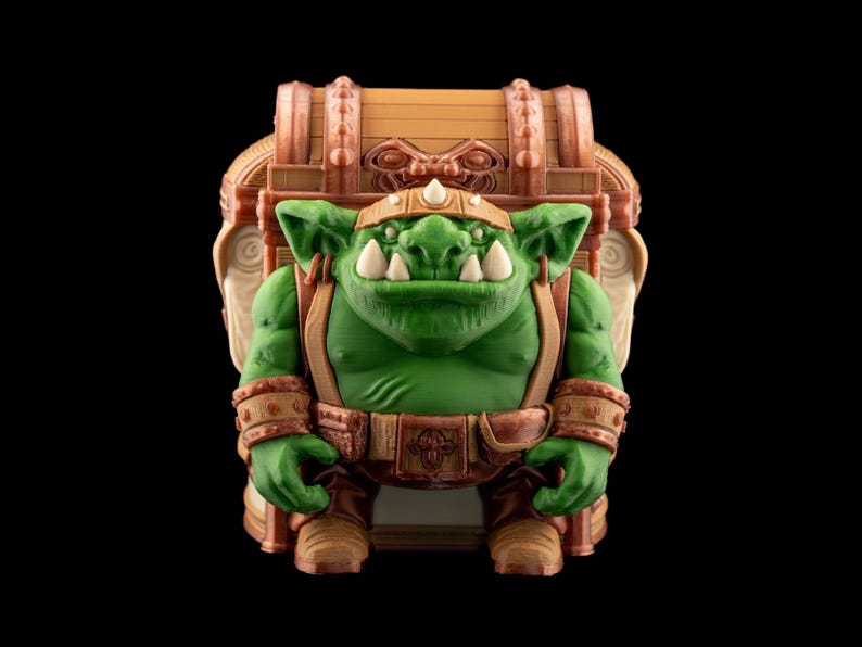 May include: A green goblin figure with large white teeth, wearing a brown belt, boots, and armbands. The goblin is carrying a treasure chest on its back. The chest is brown with copper-colored accents. The background is black.
