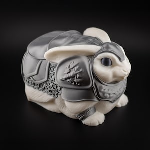 May include: A white rabbit figurine wearing gray armor. The armor covers the rabbit's back, shoulders, and forelegs. The armor has a detailed design, including chainmail and decorative elements. The rabbit has large, dark eyes and upright ears.