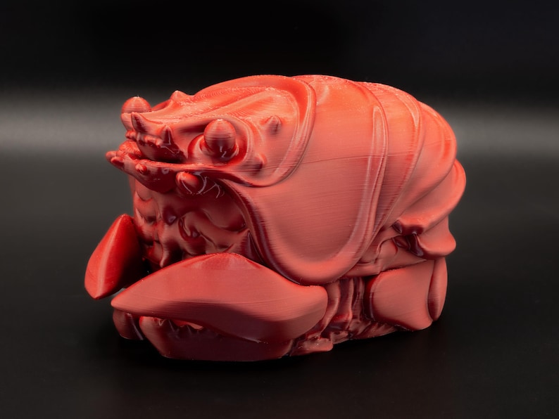 K&ouml;nnte beinhalten: Eine korallenfarbene, 3D-gedruckte Hummer-Skulptur. Der Krebs ist mit strukturierten Oberfl&auml;chen und markanten Scheren detailliert. Die Skulptur steht vor einem dunklen Hintergrund, der ihre Form hervorhebt.