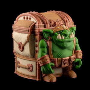 May include: A fantasy-themed figurine of a green orc character standing next to a treasure chest. The orc has large teeth, a brown leather helmet, and matching accessories. The chest is brown and beige with decorative details.