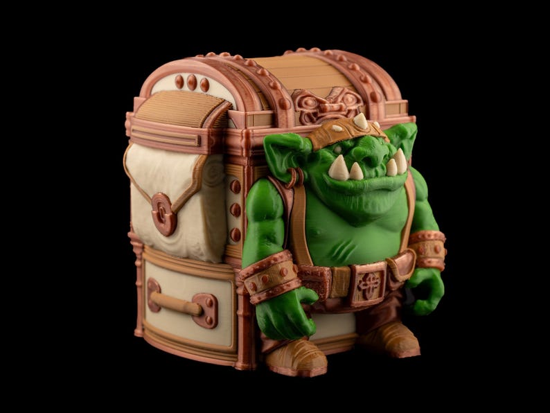 May include: A fantasy-themed figurine of a green orc character standing next to a treasure chest. The orc has large teeth, a brown leather helmet, and matching accessories. The chest is brown and beige with decorative details.