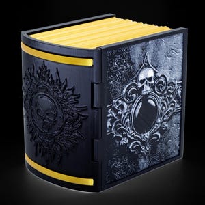May include: A black box with a gold trim and a skull design on the front. The box is closed and has a book-like appearance.