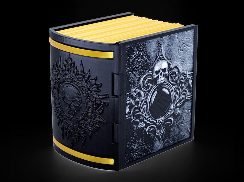 May include: A black box with a gold trim and a skull design on the front. The box is closed and has a book-like appearance.