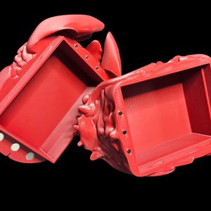 K&ouml;nnte beinhalten: Eine leuchtend rote, hummerf&ouml;rmige Box, die sich &ouml;ffnet, um einen rechteckigen Innenraum freizulegen. Die Box besteht aus einem festen Material mit strukturierter Oberfl&auml;che. Die Box ist ge&ouml;ffnet und zeigt das Innere. Sie hat einen Magnetverschluss. Der Hintergrund ist schwarz.