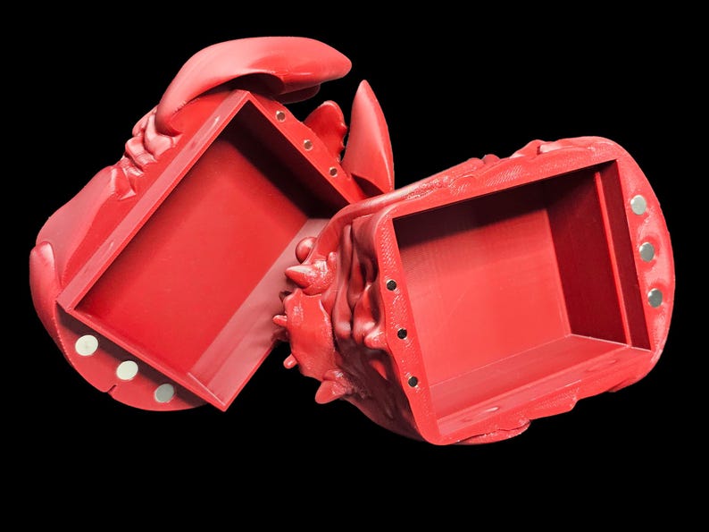 K&ouml;nnte beinhalten: Eine leuchtend rote, hummerf&ouml;rmige Box, die sich &ouml;ffnet, um einen rechteckigen Innenraum freizulegen. Die Box besteht aus einem festen Material mit strukturierter Oberfl&auml;che. Die Box ist ge&ouml;ffnet und zeigt das Innere. Sie hat einen Magnetverschluss. Der Hintergrund ist schwarz.