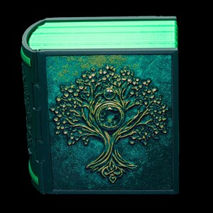 May include: A teal green book with a gold embossed tree design on the cover. The book is closed and the pages are visible, glowing green.