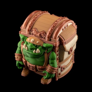 May include: A fantasy-themed decorative chest with a green goblin figure integrated into its design. The chest is brown and beige with bronze accents. The goblin has a green complexion, large teeth, and is wearing a brown helmet and belt.