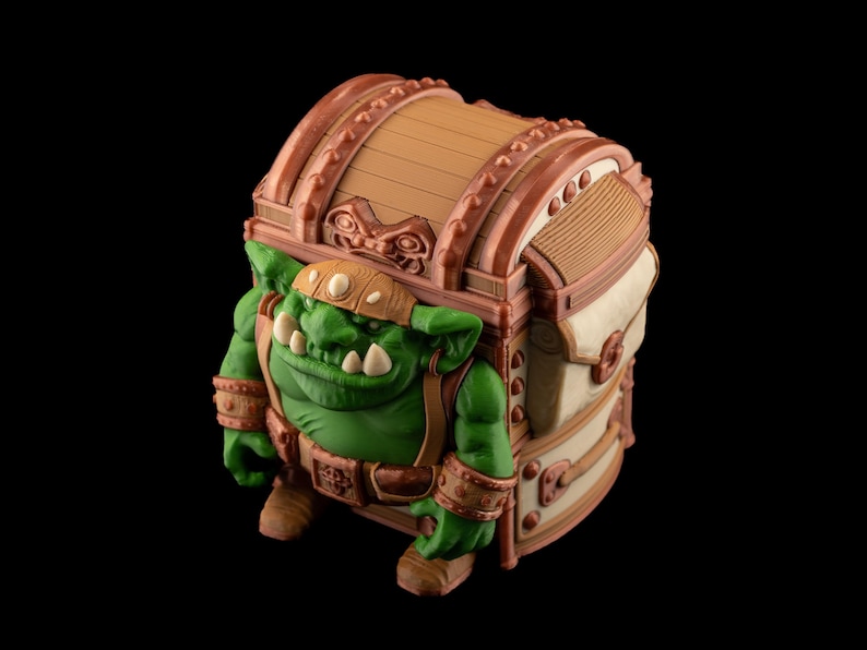 May include: A fantasy-themed decorative chest with a green goblin figure integrated into its design. The chest is brown and beige with bronze accents. The goblin has a green complexion, large teeth, and is wearing a brown helmet and belt.