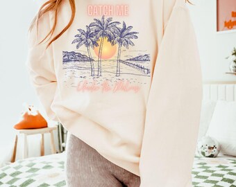 Może przedstawiać: Różowa bluza z nadrukiem palm i zachodu słońca. Tekst "CATCH ME" znajduje się nad nadrukiem, a tekst "Under the Palms" pod nadrukiem.