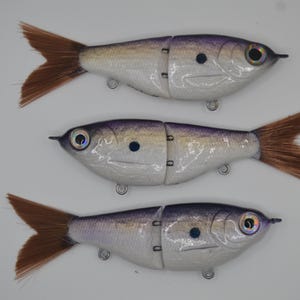 May include: Three artificial fish lures with segmented bodies, designed to mimic baitfish. Each lure features a silver body with purple accents, a brown tail, and realistic eye details. The lures are suspended with small metal hooks.