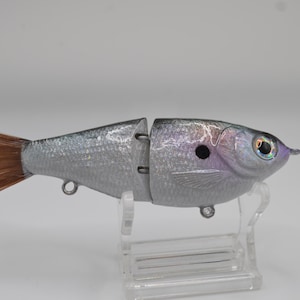 May include: A silver and gray articulated fishing lure shaped like a fish. The lure has a brown tail and a detailed head with a black eye and iridescent accents. It is designed to mimic a baitfish and is mounted on a clear stand.