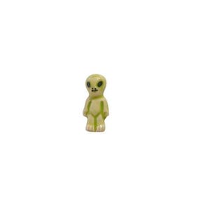 May include: A small, ceramic alien figurine in shades of pale green and yellow. The alien has large, black eyes and a small, smiling mouth. The figurine is standing upright with arms at its sides.