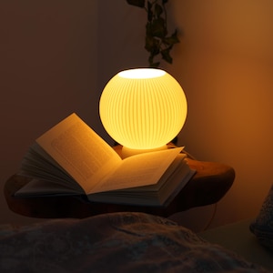Könnte beinhalten: Eine leuchtende, kugelförmige Lampe mit geripptem Design steht auf einem offenen Buch auf einer Holzoberfläche. Das warme Licht erhellt die Seiten und die Umgebung und schafft eine gemütliche Atmosphäre. Im Hintergrund ist ein gemustertes Kissen zu sehen.