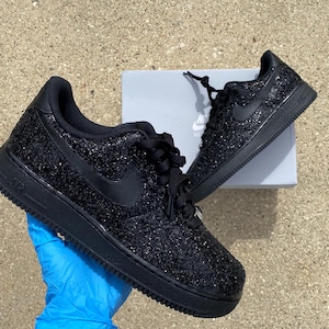 May include: Pair of black glitter-covered Nike Air Force 1 trainers. The shoes feature a black sole, laces, and the Nike swoosh logo. They are presented on a light grey surface, with a white Nike box visible.